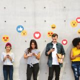 6 junge Menschen stehen vor einer Mauer. Sie schauen auf ihre Handys. Über den Handys sind Social Media Icons wie Emojis, Herzen und Daumen hoch zusehen.